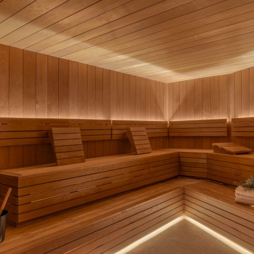A wooden sauna interior with tiled floor, benches and backrests, a bucket, towels, and fresh herbs in the corner.