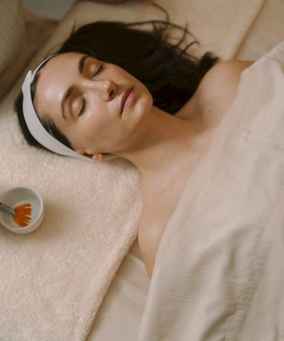 A woman lying in bed, eyes closed, headband, covered with a blanket, receiving a facial massage with a brush on her face.