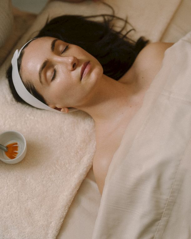 A woman lying in bed, eyes closed, headband, covered with a blanket, receiving a facial massage with a brush on her face.
