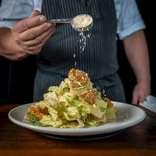 A chef sprinkles grated cheese or sauce over a loaded plate of creamy salad greens and croutons it&rsquo;s being plated.