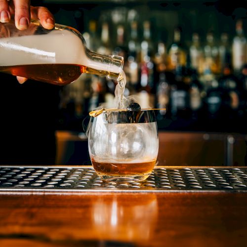 A bartender pours a splash of whiskey into a glass over ice, with bottles lined up in the blurred bar backdrop, shot at bar height.