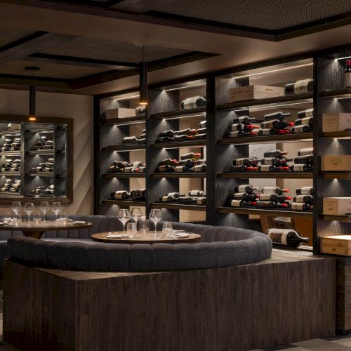 Luxury wine cellar with rows of bottled red wine and a tasting table with glasses beneath dark shelves.