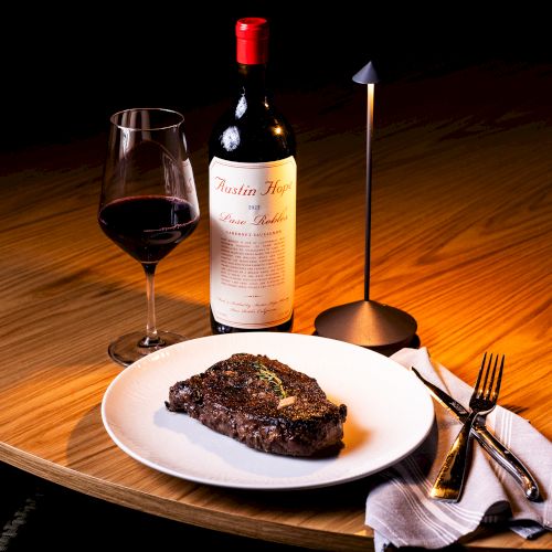 A candlelit table with a steak on a plate, a glass of red wine, a bottle, and a lit candle in the background.