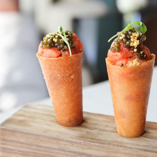 Two small, conical, orange-colored hors d'oeuvres stand upright on a wooden block, topped with greens and chopped nuts.