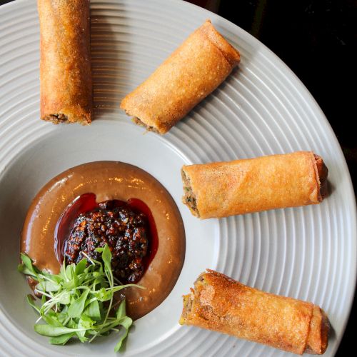 Crispy golden spring rolls arranged around a bowl of dipping sauce with a garnish on a white plate, four rolls visible.