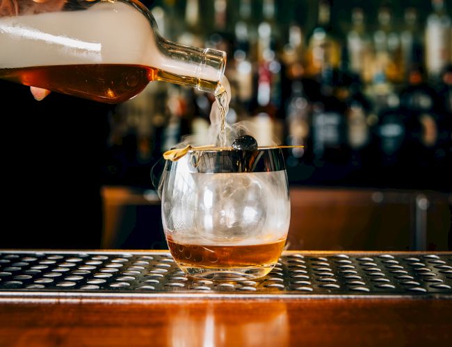 A bartender pours whiskey over ice into a glass, as bottles line the bar in the background, creating a moody, classy ambiance.