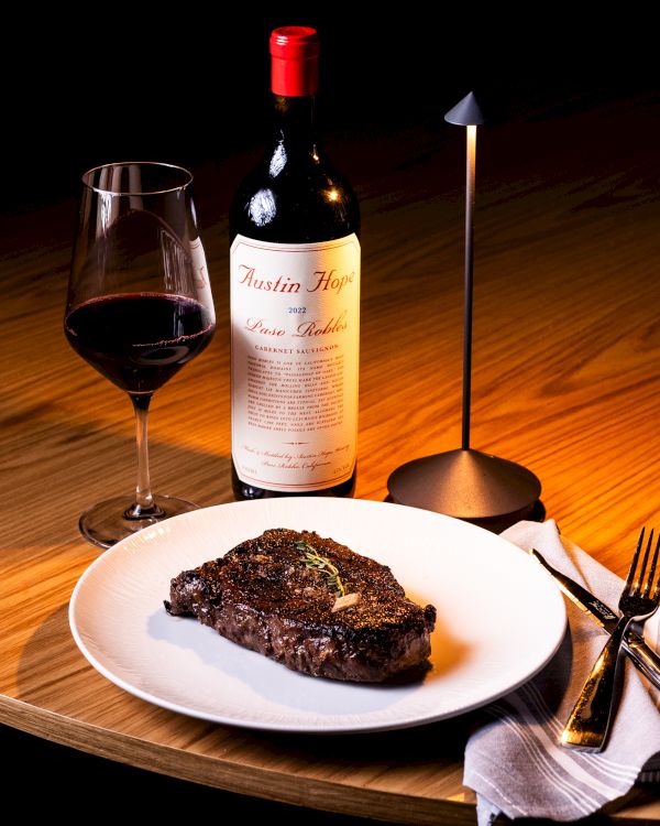A dinner setup: a glass of red wine, a bottle, a plate with steak, a candle, and cutlery on a wooden table.