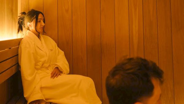 A woman in a yellow robe sits in a wooden sauna while a man faces her, both relaxing in warm light.