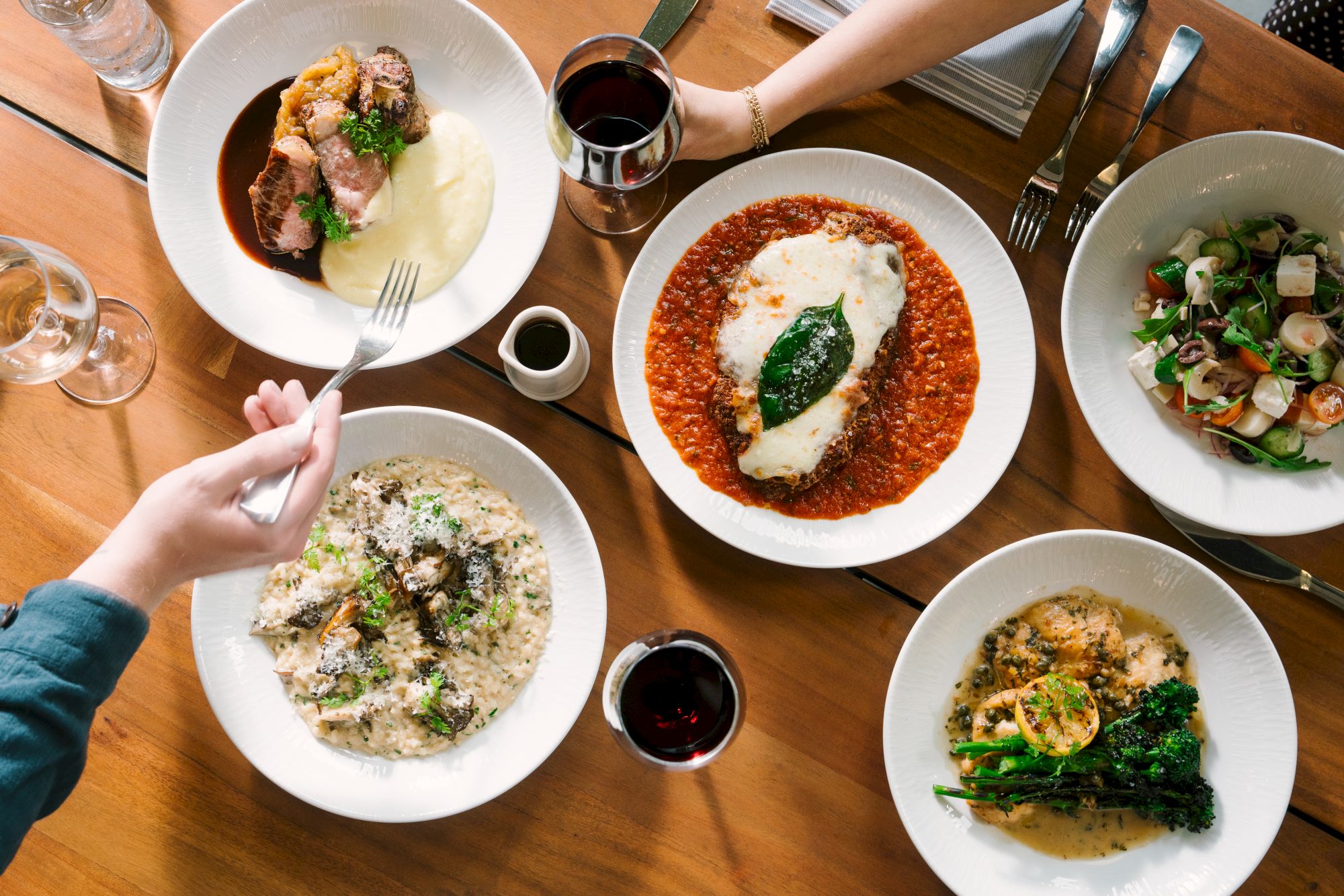 A table of plated dishes&mdash;lasagna with red sauce, creamy pasta, roast meat, a fresh salad, and glasses of wine&mdash;ready to eat.