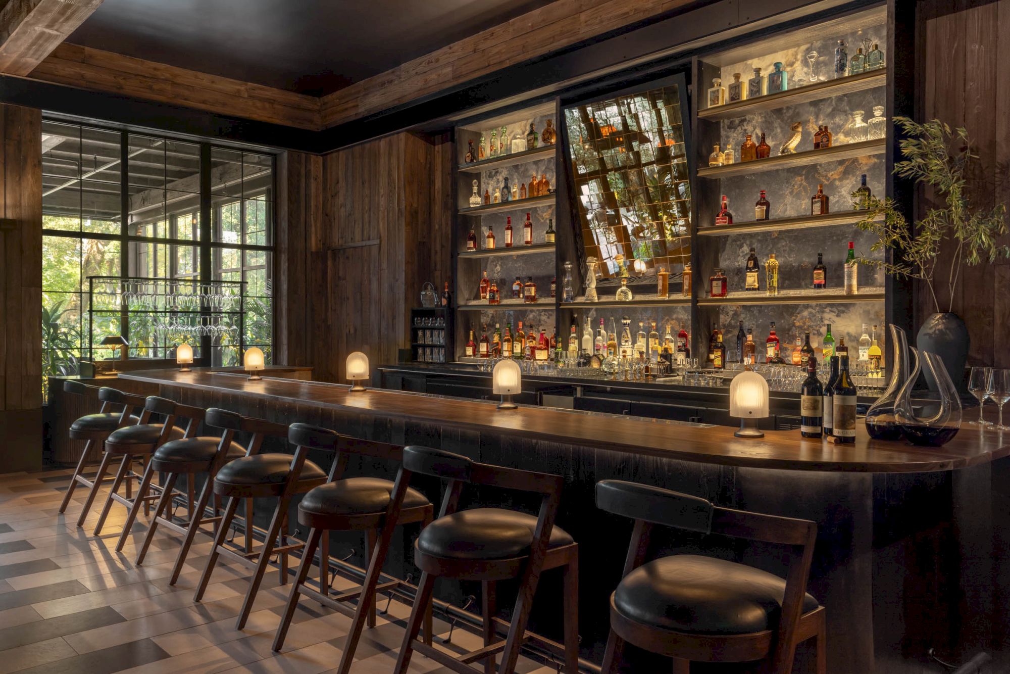A cozy wooden cocktail bar with glass shelves of liquor bottles, pendant lights, and high stools by large windows.