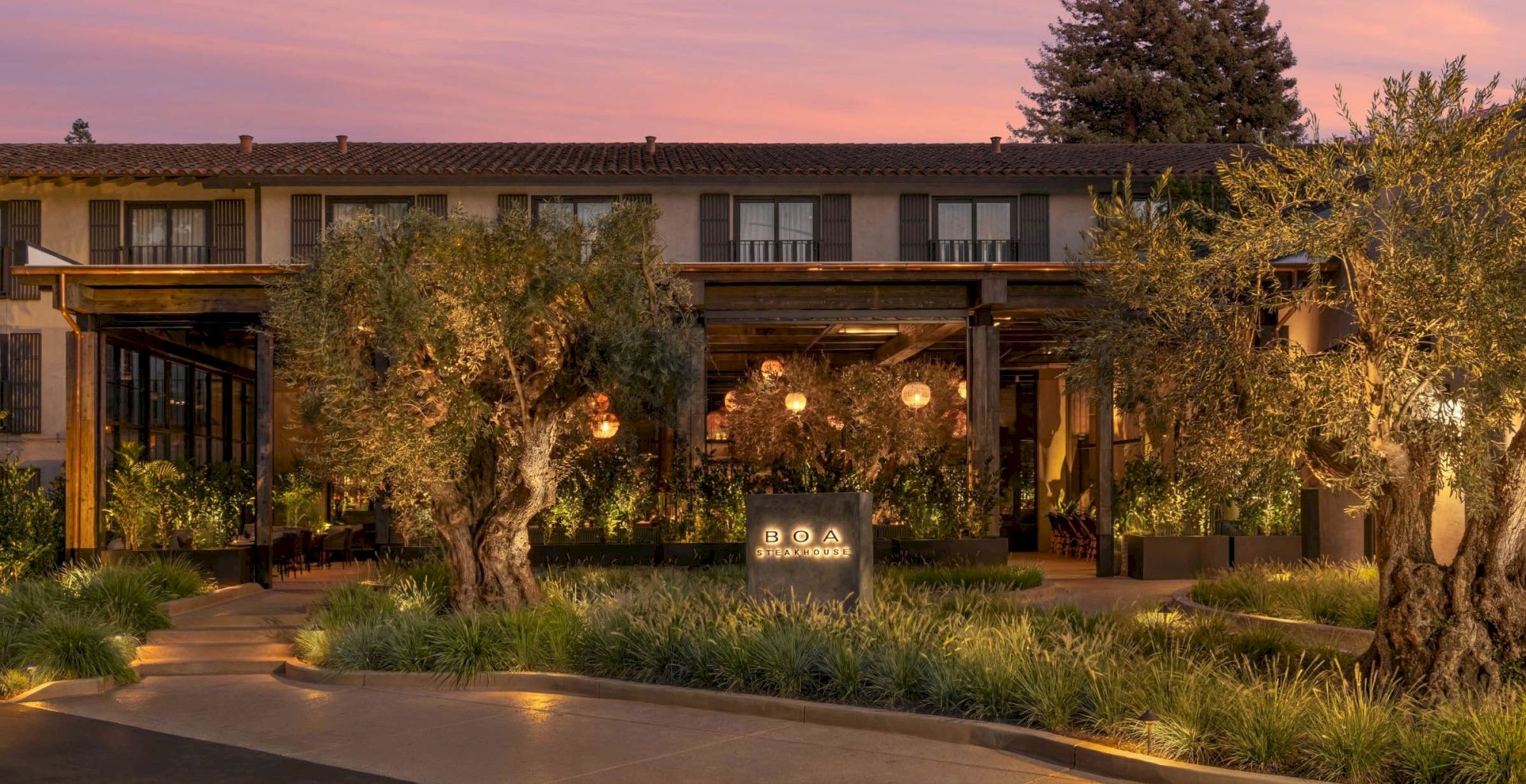 Evening view of a hotel exterior and patio with glowing lights, olive trees, and a sign reading BOA Steakhouse beneath a pink sky.