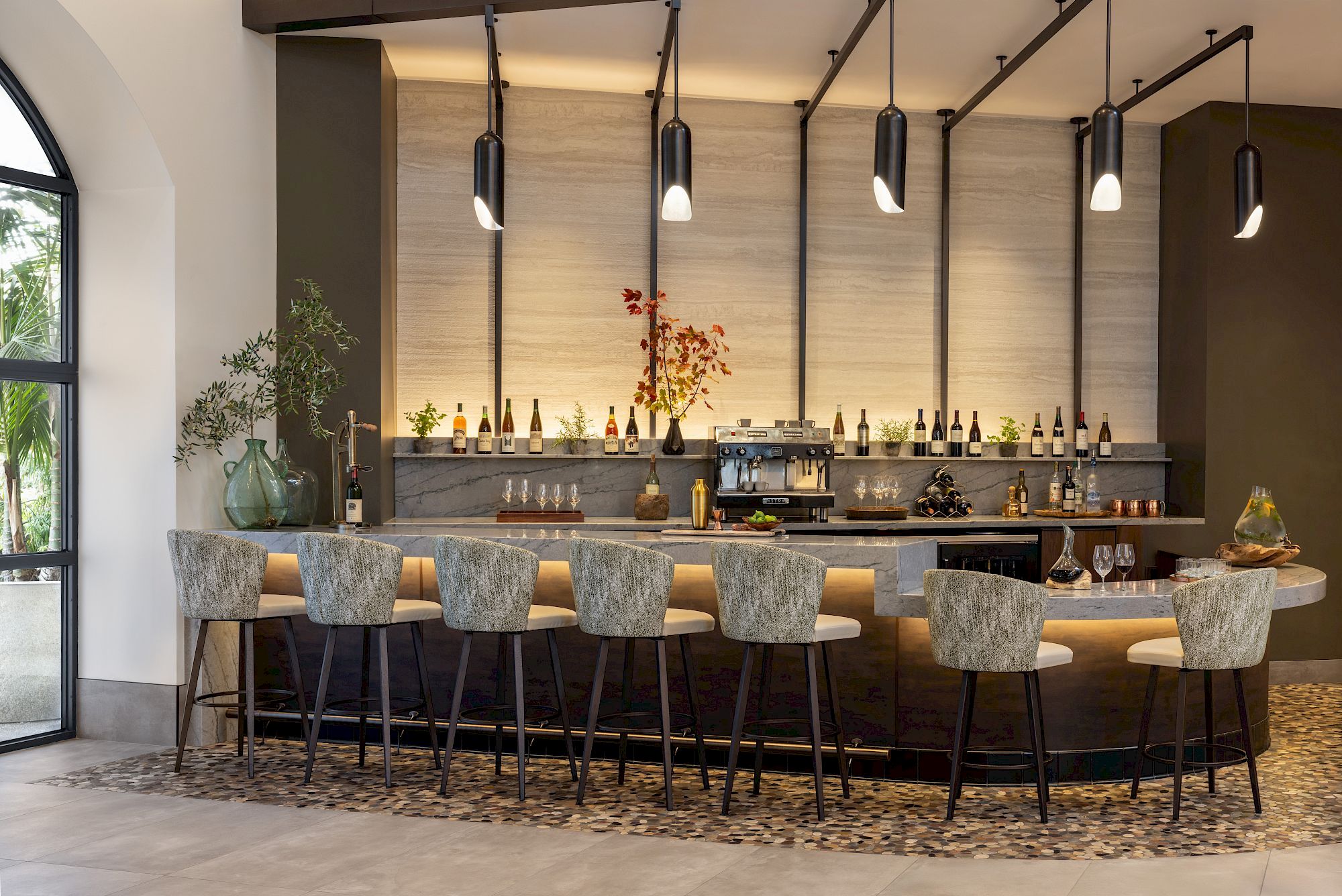 A modern bar with pendant lights, bottles, a coffee/espresso station, and high stools arranged along a stone counter.