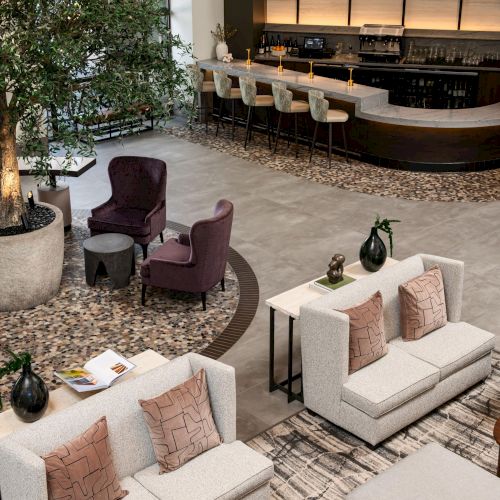 A modern lounge with beige sofas, a purple armchair, a curved bar with pendant lights, and indoor greenery fills the scene.