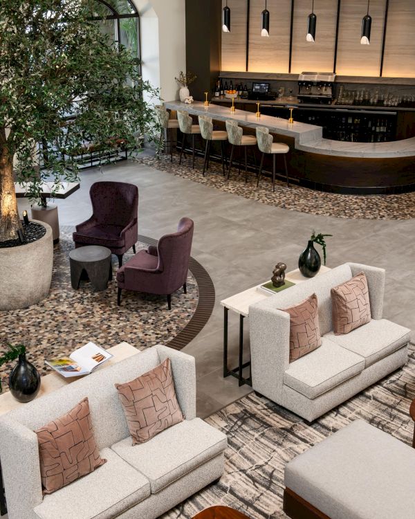 A modern lounge with beige sofas, a purple armchair, a curved bar with pendant lights, and indoor greenery fills the scene.
