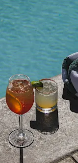Two cocktails with ice and lime sit on a poolside ledge, with a folded towel nearby in sunny blue water.
