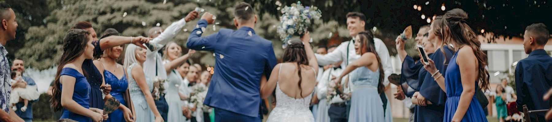 An outdoor wedding celebration as the groom carries the bride, while guests cheer and toss confetti under string lights.