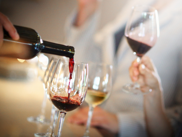 A hand pours red wine from a bottle into a glass while other guests toast in the background.