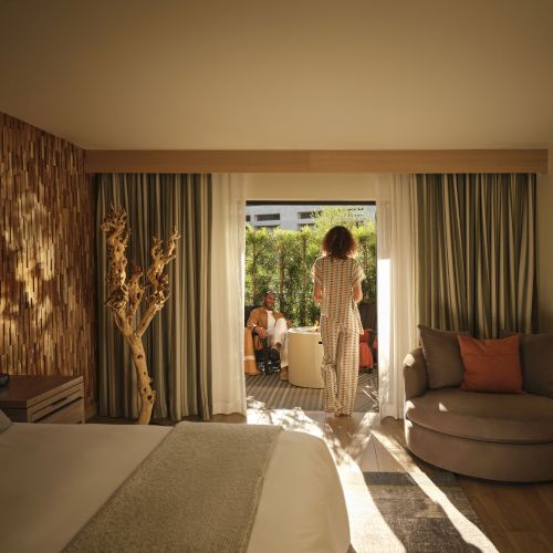 A bright hotel lounge with a couch and curtains, a sculptural wood wall, and a woman greeting a man outside through open doors.