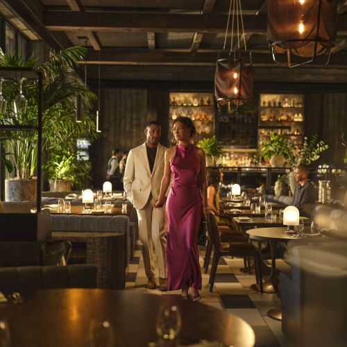 A stylish couple walks through a candlelit restaurant with plants, hanging lights, and a bar in the background.