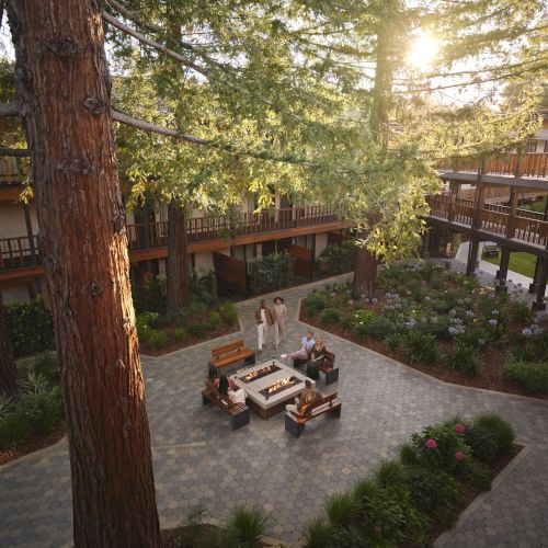 Sunset courtyard with towering trees, wooden balconies, gardens, and people relaxing beside a lit firepit in a brick plaza.