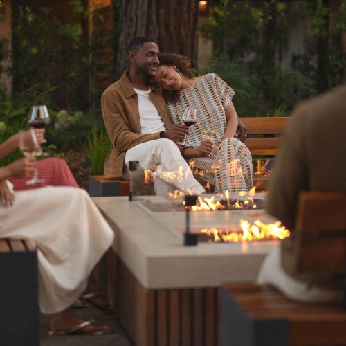 A couple smiles and cheers over wine at an outdoor fire pit, with friends chatting nearby.