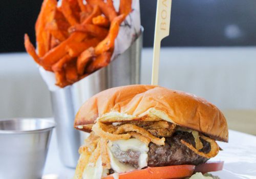 A juicy burger with melted cheese and veggies sits on a plate beside a cup of crispy orange fries, ready to eat.